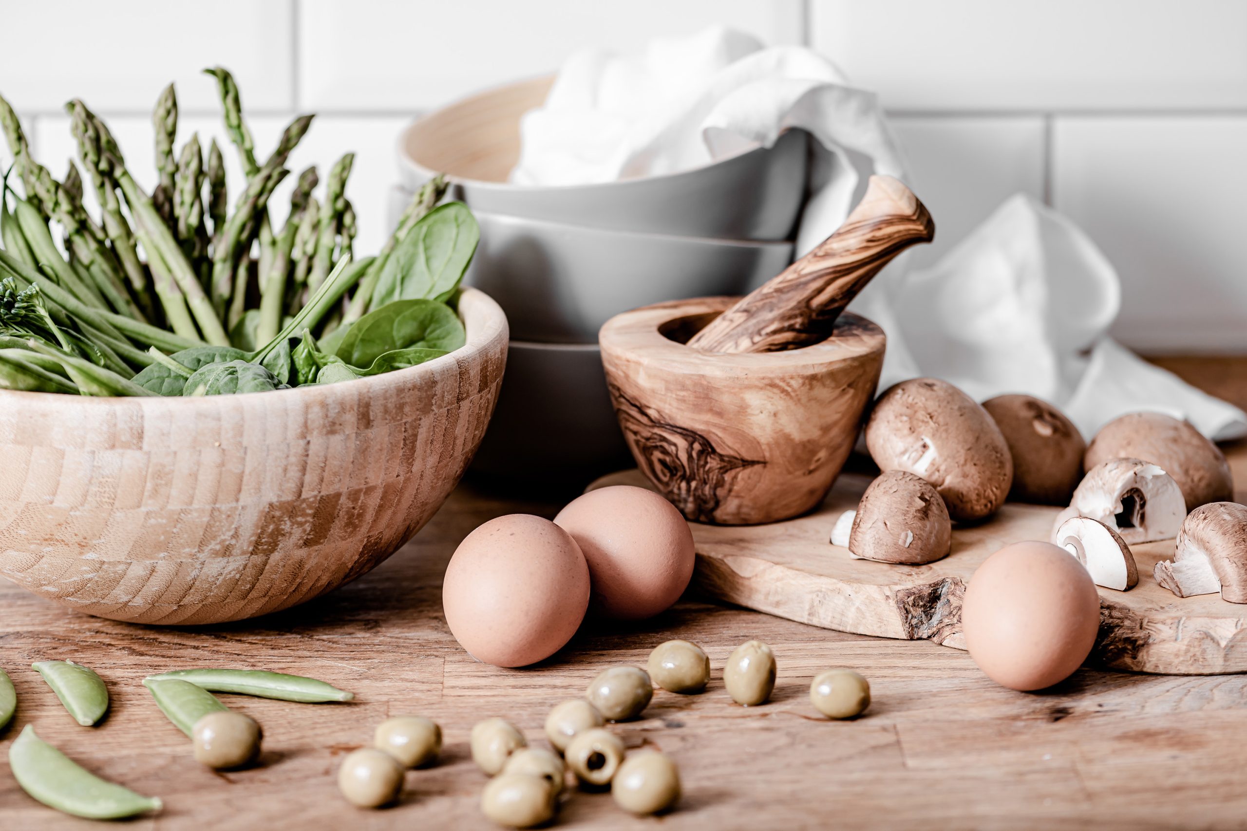 A wooden bowl filled with asparagus and spinach sits next to a cutting board with brown mushrooms, eggs, and green olives. Nearby are stacked bowls, a mortar and pestle, and a white cloth on a wooden counter.