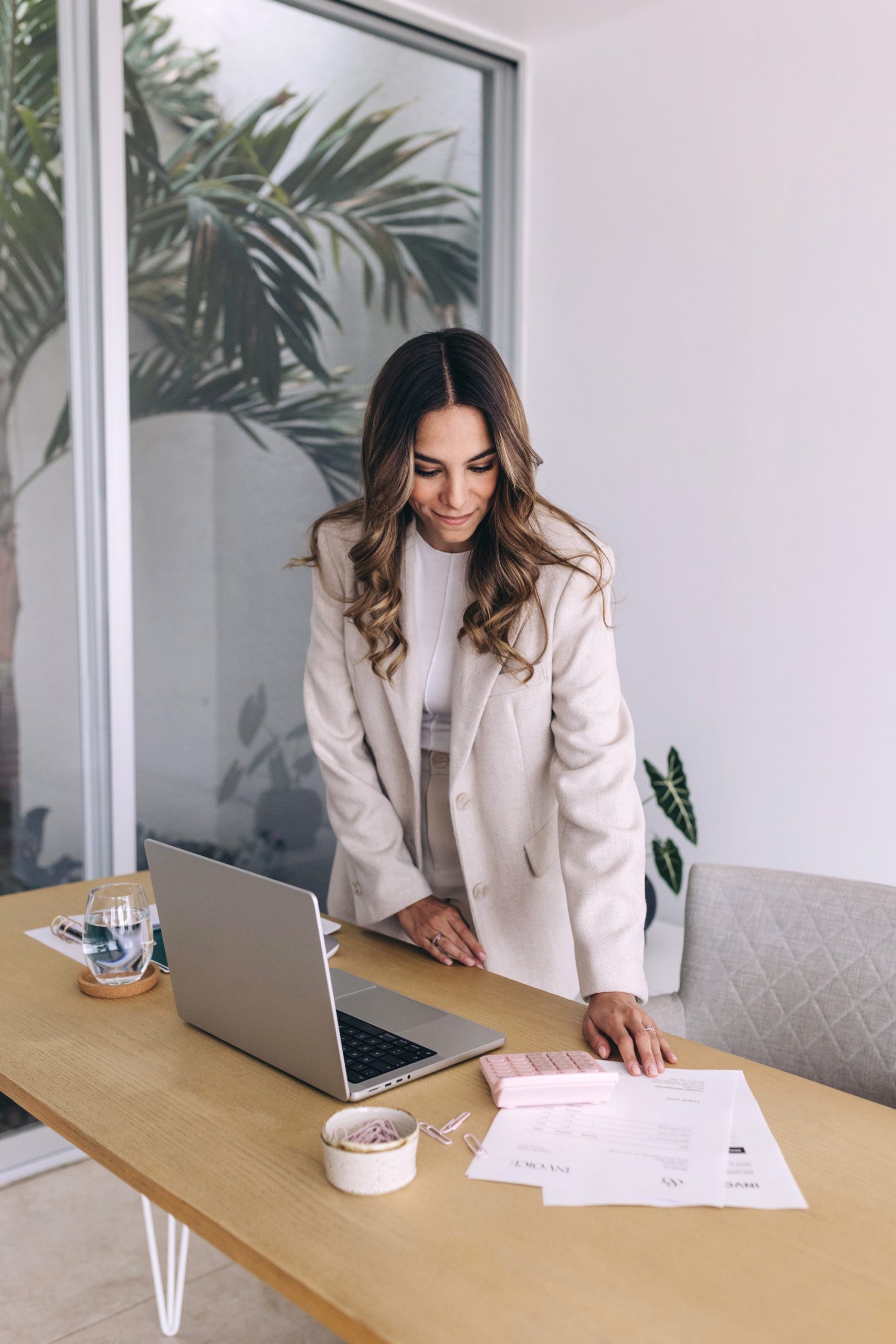 A woman in a light-colored blazer stands at a desk with a laptop, papers, and a glass of water. She looks down, smiling slightly, in a modern, bright room with large windows and indoor plants.
