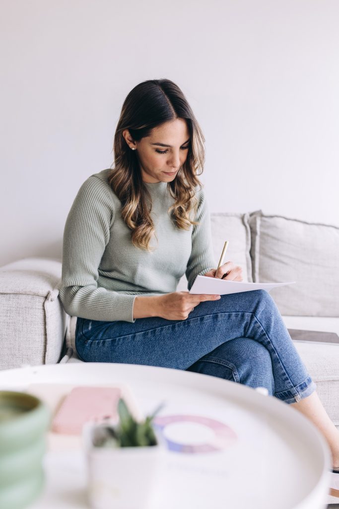A woman with long hair sits on a light-colored sofa, wearing a green sweater and jeans, writing on a notepad with a pen. A table with a plant and papers is in the foreground.