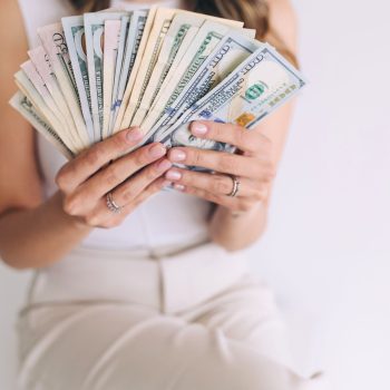 A woman in light-colored clothing smiles while holding a fanned-out stack of U.S. dollar bills in both hands, showing her manicured nails and several rings.