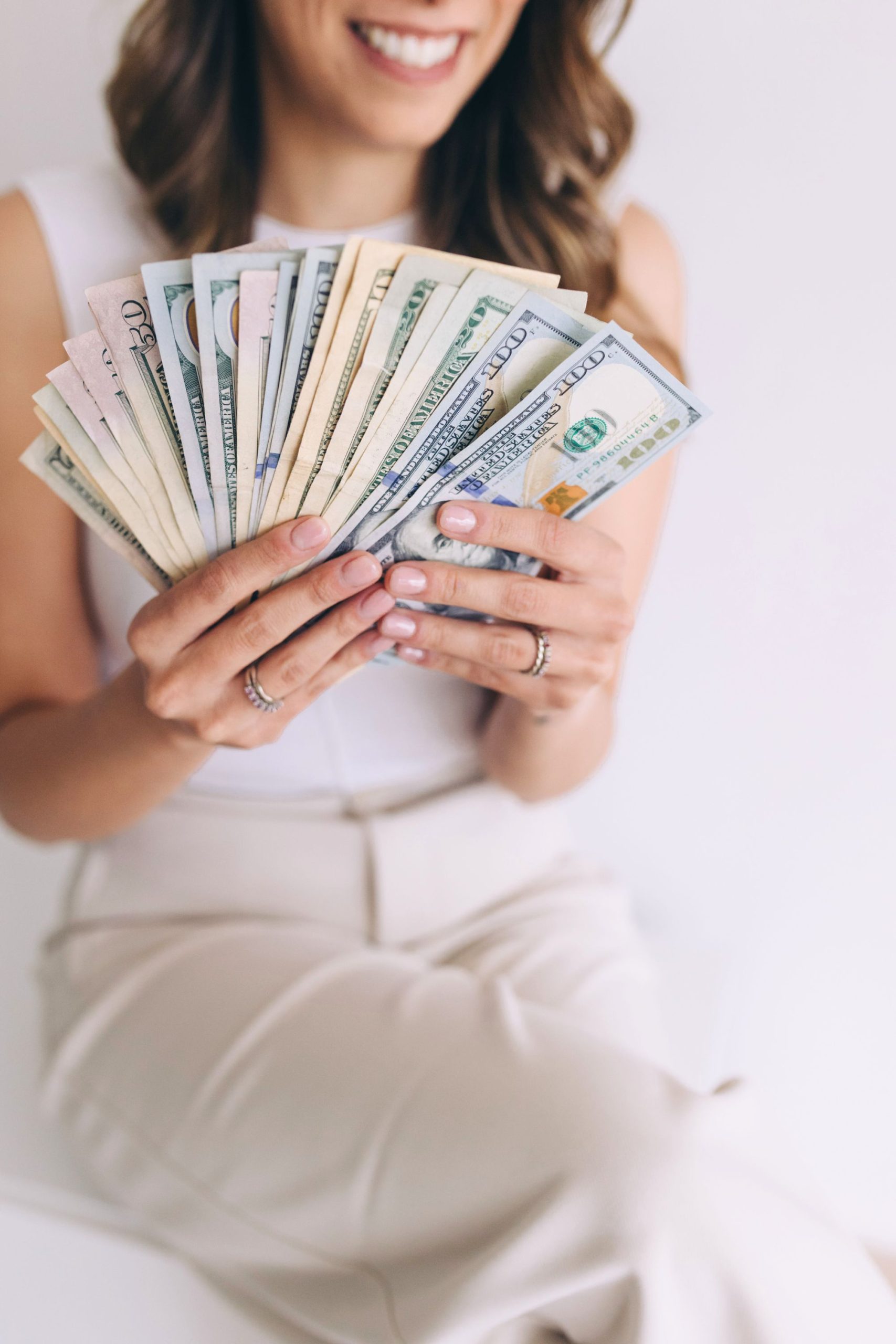 A woman in light-colored clothing smiles while holding a fanned-out stack of U.S. dollar bills in both hands, showing her manicured nails and several rings.
