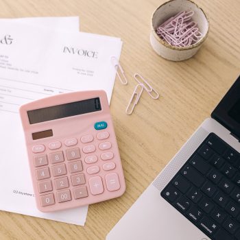 A pink calculator sits on a desk next to an invoice, a laptop keyboard, and a bowl of pink paper clips. Some paper clips are scattered on the wooden surface.