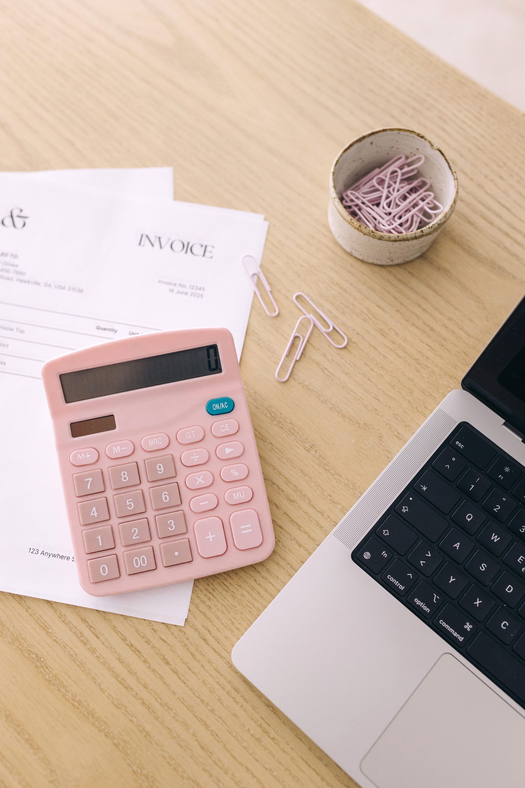 A pink calculator sits on a desk next to an invoice, a laptop keyboard, and a bowl of pink paper clips. Some paper clips are scattered on the wooden surface.
