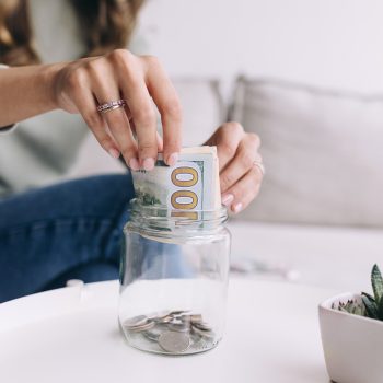 A woman places a $100 bill into a glass jar partially filled with coins, sitting on a white table with a small potted plant nearby.