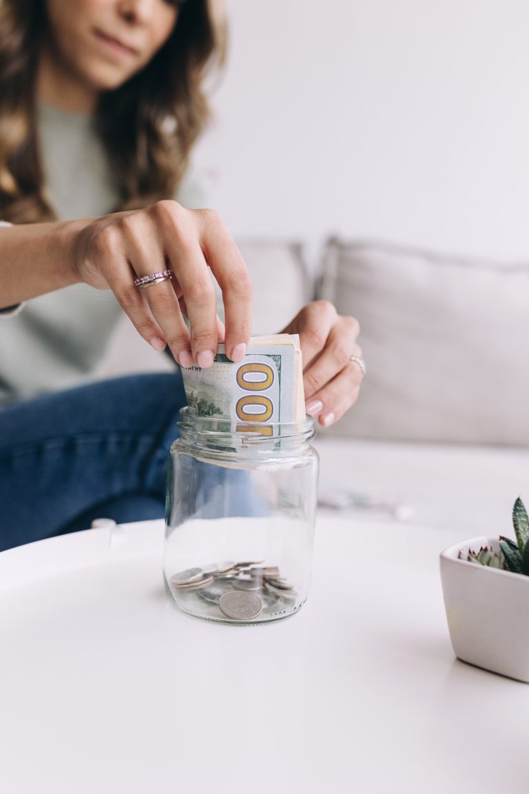 A woman places a $100 bill into a glass jar partially filled with coins, sitting on a white table with a small potted plant nearby.