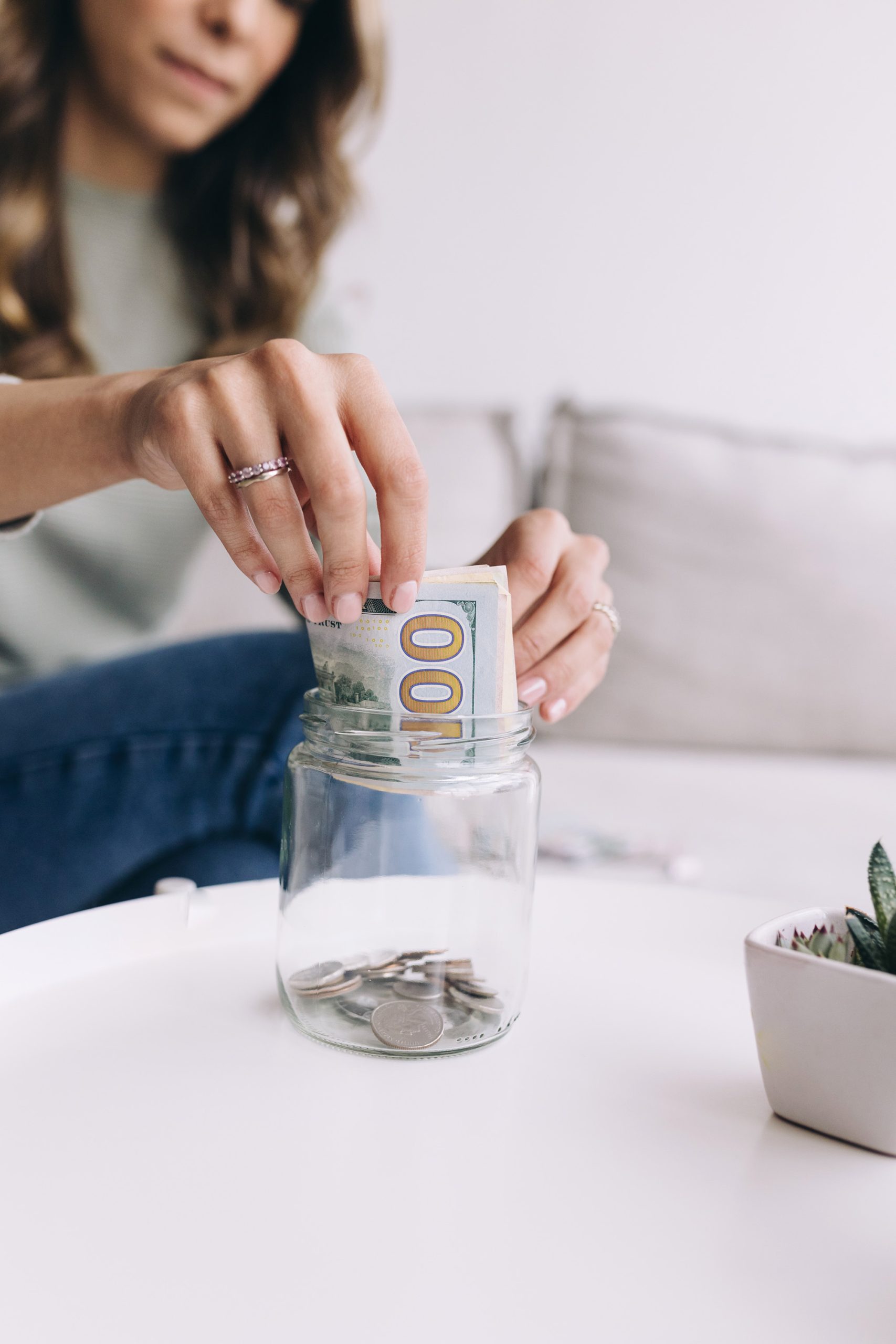 A woman places a $100 bill into a glass jar partially filled with coins, sitting on a white table with a small potted plant nearby.