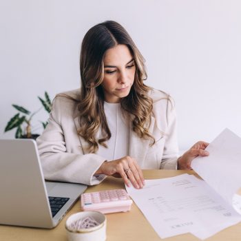 A woman sits at a desk with a laptop, calculator, and documents, reviewing paperwork. She appears focused and is wearing a light-colored blazer. There is a potted plant and a bowl of paperclips on the desk.