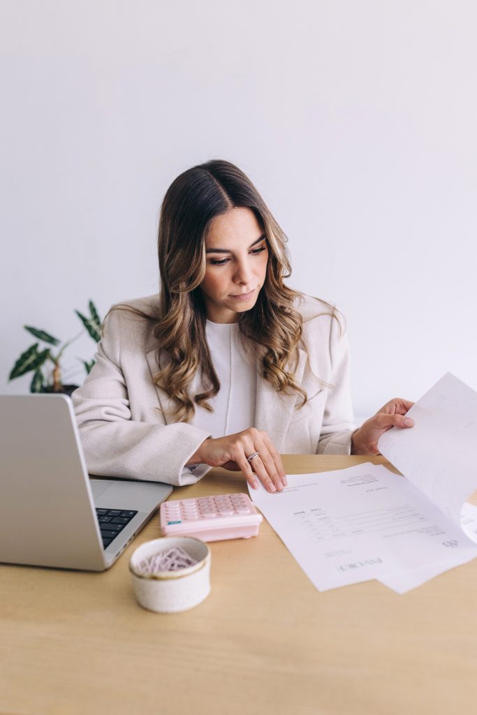 A woman sits at a desk with a laptop, calculator, and documents, reviewing paperwork. She appears focused and is wearing a light-colored blazer. There is a potted plant and a bowl of paperclips on the desk.