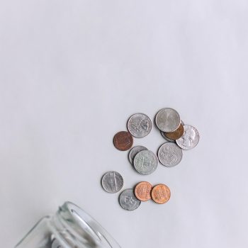 A glass jar tipped over on a light surface with several coins, including quarters, dimes, nickels, and pennies, spilling out.