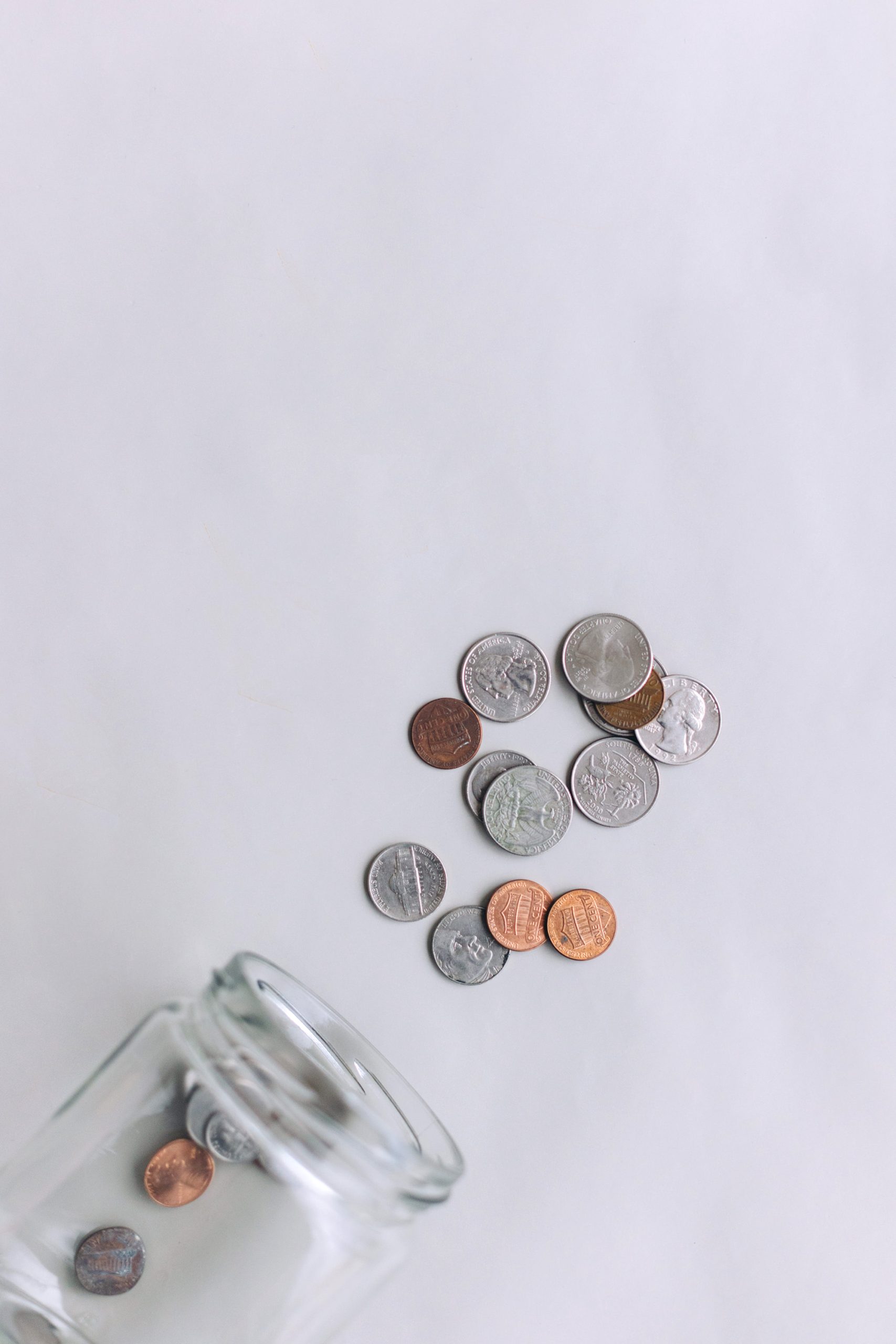 A glass jar tipped over on a light surface with several coins, including quarters, dimes, nickels, and pennies, spilling out.