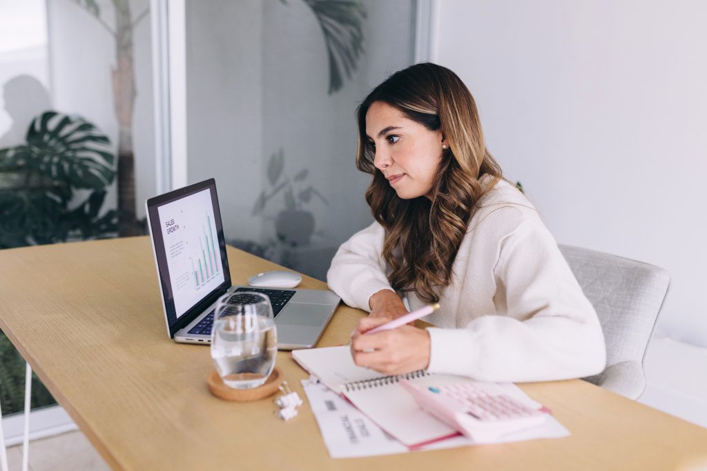 A woman sits at a desk working on a laptop with graphs on the screen. She writes in a notebook, with a calculator, documents, and a glass of water beside her. She appears focused in a home office setting.