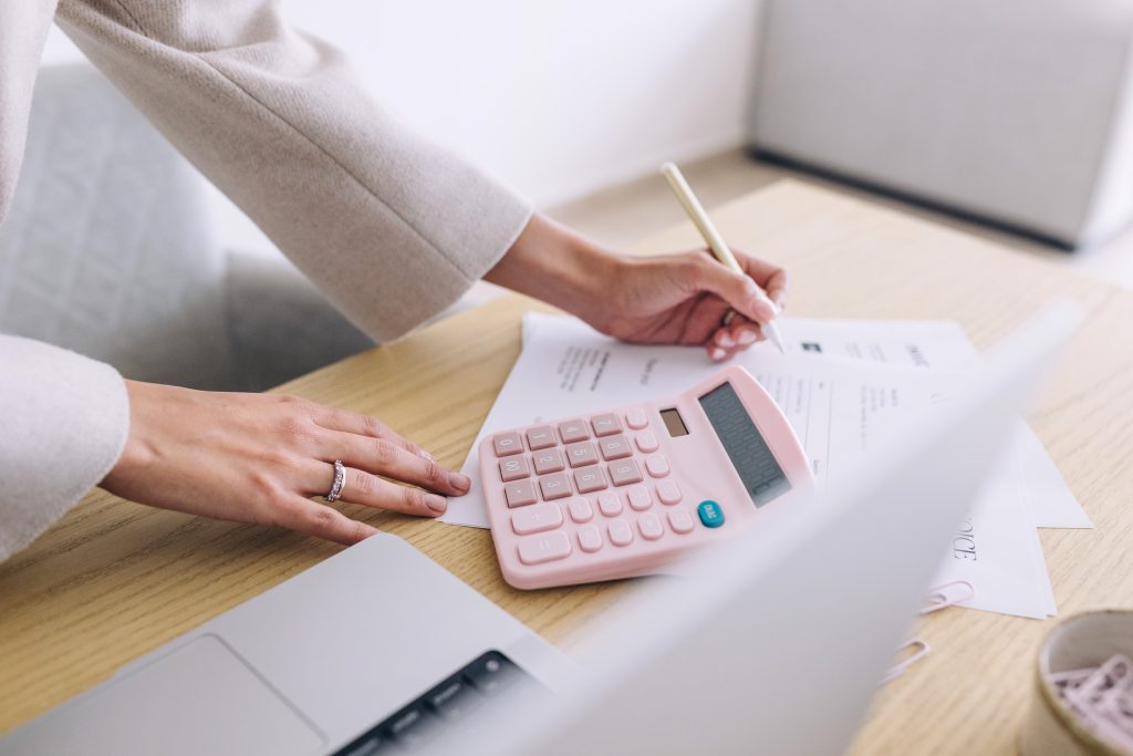 Close-up of a person’s hands using a pink calculator and writing on documents at a desk, with a laptop nearby. The scene suggests working on finances, budgeting, or administrative tasks.