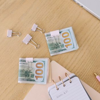 Stacks of 100-dollar bills clipped with binder clips on a wooden desk, alongside a spiral notepad, folders, binder clips, a pencil, and part of a laptop.