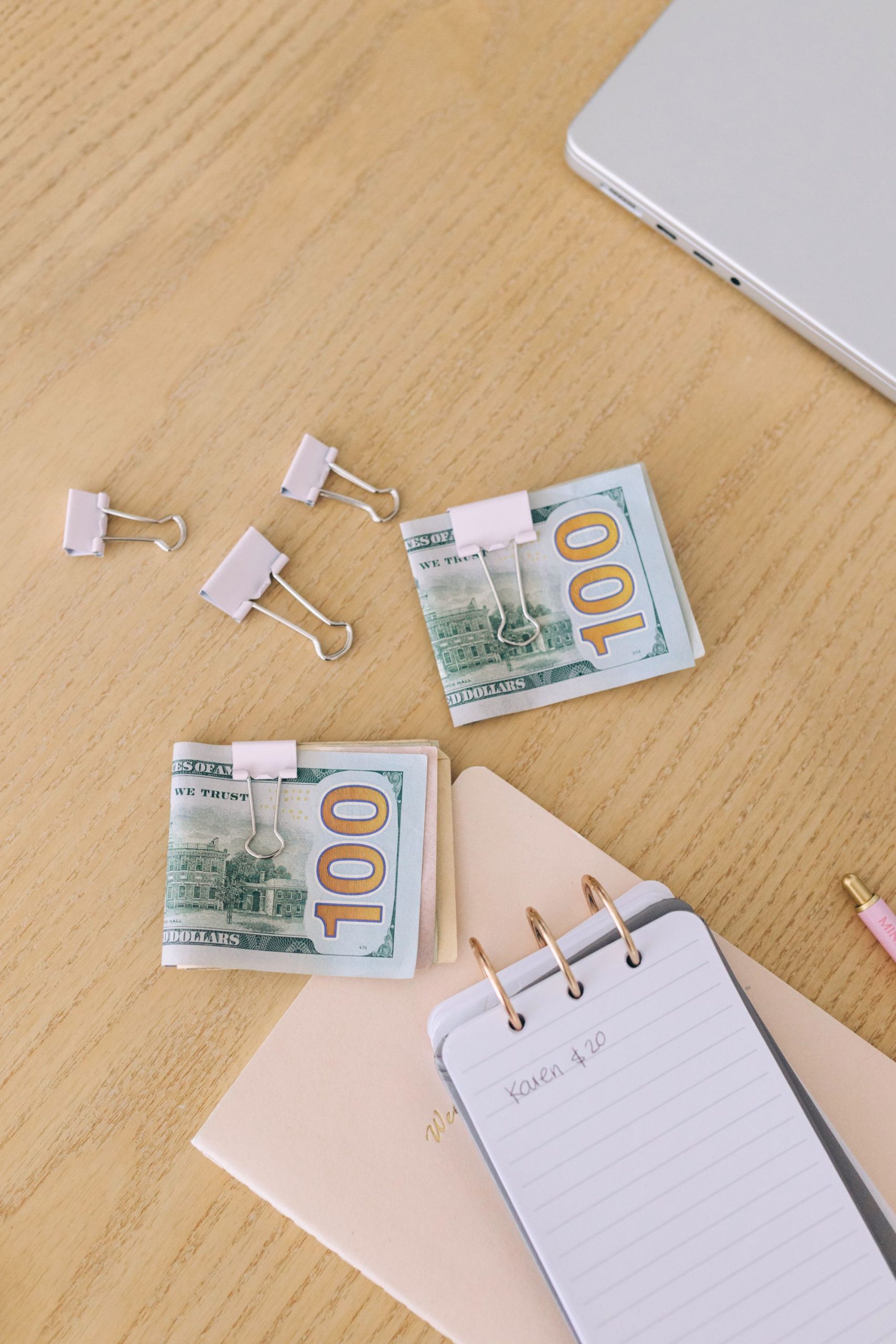 Stacks of 100-dollar bills clipped with binder clips on a wooden desk, alongside a spiral notepad, folders, binder clips, a pencil, and part of a laptop.