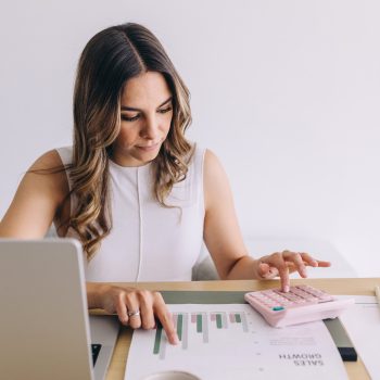 A woman sits at a desk with a laptop, using a pink calculator and pointing at a printed sales growth chart. A small bowl with paper clips is also on the desk. She appears focused on her work.