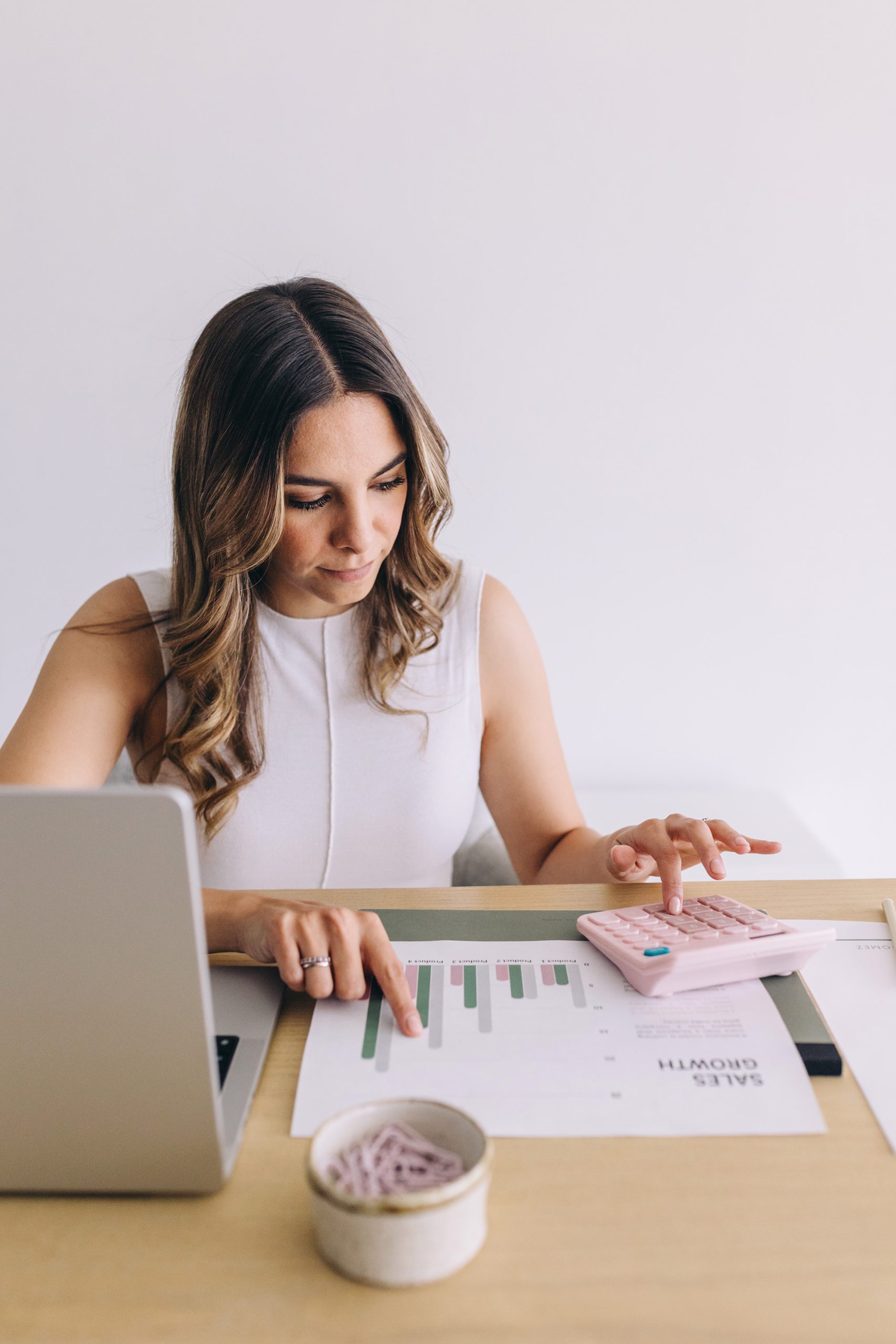 A woman sits at a desk with a laptop, using a pink calculator and pointing at a printed sales growth chart. A small bowl with paper clips is also on the desk. She appears focused on her work.