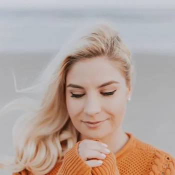 A woman with long blonde hair, wearing an orange sweater, stands outdoors with her eyes closed and hands clasped together near her face, appearing peaceful and content. The background is softly blurred.