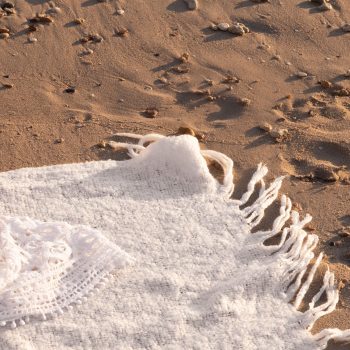 A close-up of a white fringed blanket spread on sandy beach, with small pebbles scattered nearby and intricate lace detail partly visible in warm sunlight.
