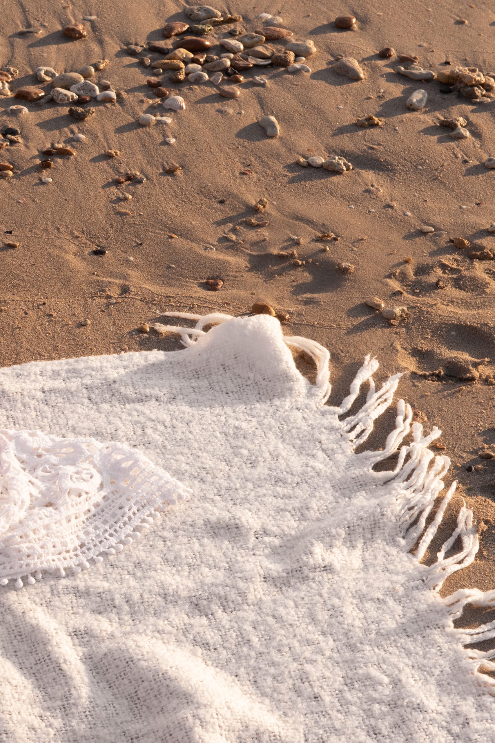 A close-up of a white fringed blanket spread on sandy beach, with small pebbles scattered nearby and intricate lace detail partly visible in warm sunlight.