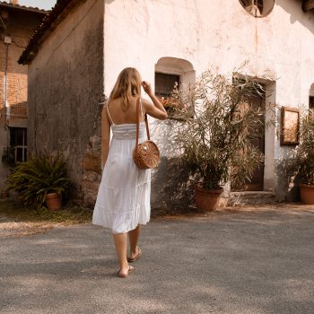A woman in a white sundress walks barefoot on a sunlit street toward an old, rustic house with potted plants and weathered walls. She carries a round, woven bag over her shoulder.