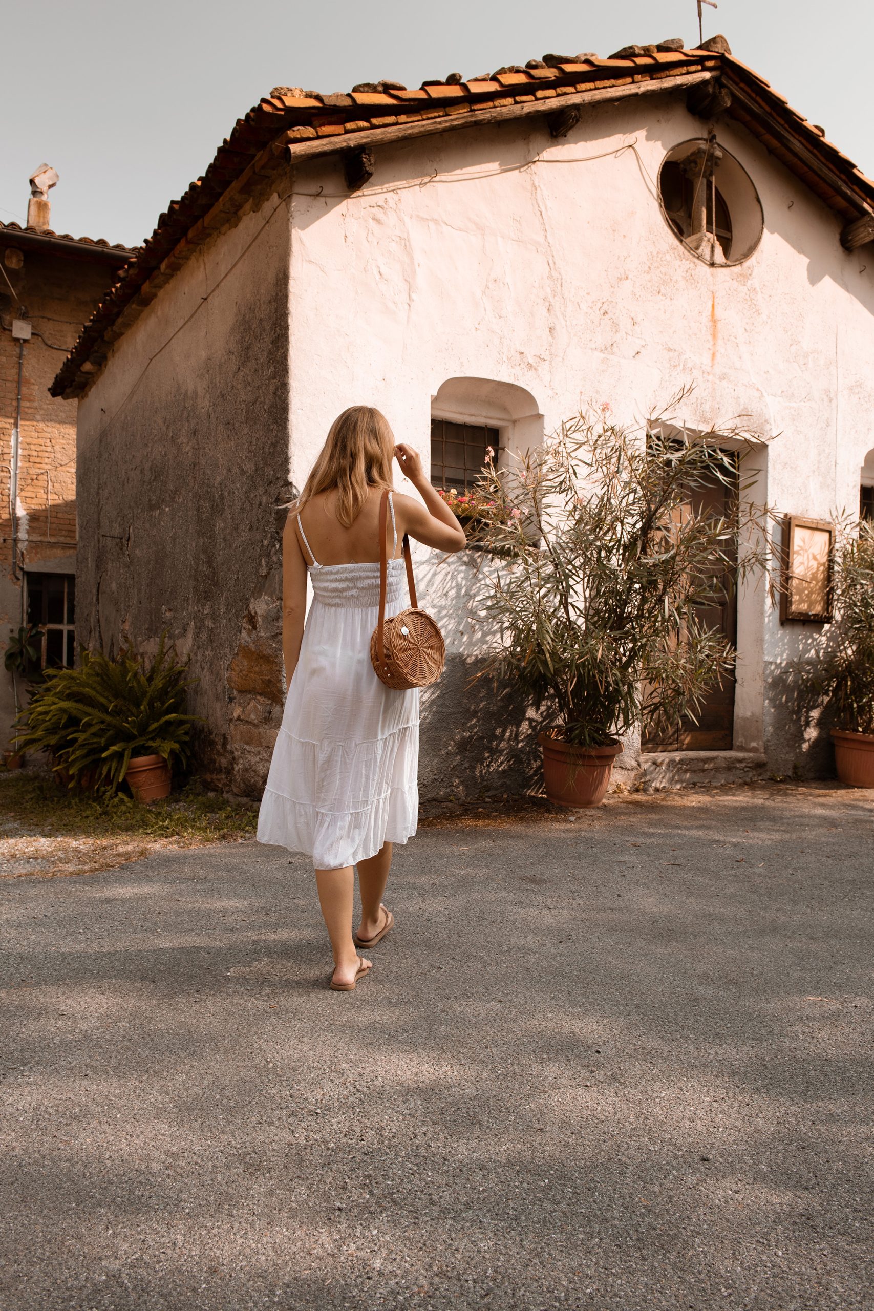A woman in a white sundress walks barefoot on a sunlit street toward an old, rustic house with potted plants and weathered walls. She carries a round, woven bag over her shoulder.
