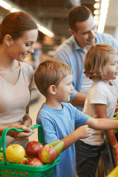 A photograph of a family of four—a mother, father, and two young children—browsing the produce section of a bustling grocery store.