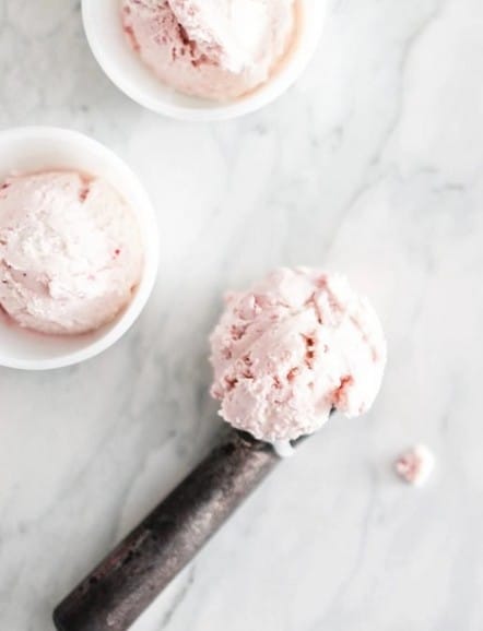 A metal ice cream scooper with a scoop of pale pink ice cream rests on a marble surface next to two small white bowls, each containing a scoop of the same ice cream.