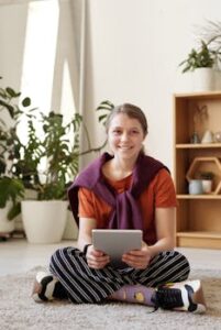 Cheerful teenager sitting on the floor using a tablet, surrounded by plants at home.
