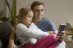 A father and daughter enjoying a relaxed moment together using a smartphone indoors.