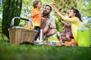 A family with children enjoying a picnic in a sunny park surrounded by greenery and trees.