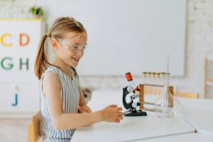 Smiling young girl exploring science with a microscope and test tubes in a classroom.