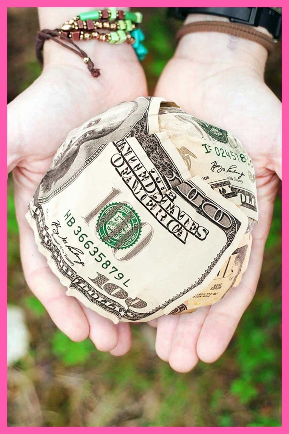 Two hands are cupped together holding a ball made of folded U.S. hundred-dollar bills, with green foliage blurred in the background and a pink border around the image.
