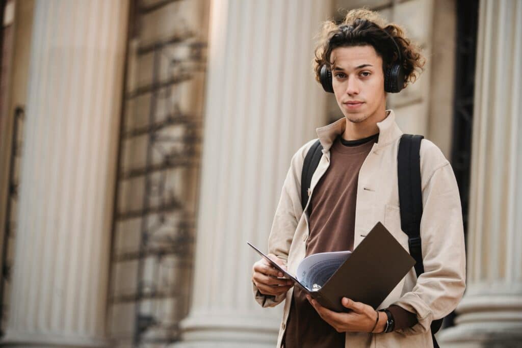 Young learner in wireless headphones in backpack standing with documents near entrance of university building