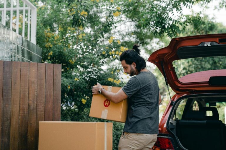 A man is loading or unloading a cardboard box from the trunk of a red car outdoors, with trees and a wooden fence in the background.