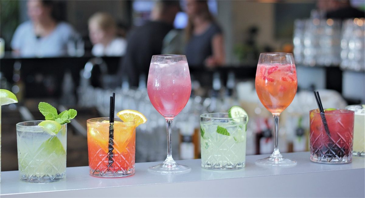 Six colorful cocktails with garnishes, including lime, lemon, mint, and berries, are lined up on a bar counter with a blurred background of people and bottles.