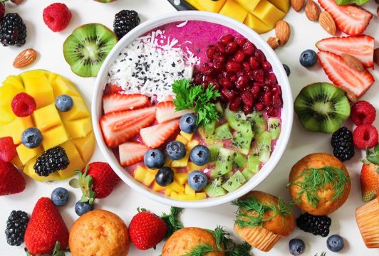 A colorful smoothie bowl topped with sliced strawberries, blueberries, kiwi, mango, pomegranate, shredded coconut, and parsley, surrounded by various fresh fruits and small muffins.