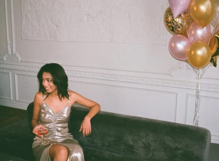 A woman in a shiny silver dress sits on a dark sofa holding a drink, smiling. Pink, gold, and confetti balloons are on her right. The background features white textured walls and decorative molding.