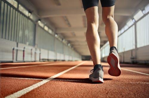 Person in athletic wear walking or running on an indoor track with hurdles in the background. The focus is on their legs and running shoes.