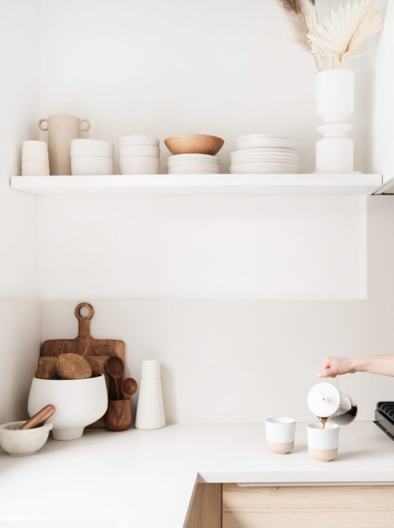 Minimalist kitchen with white walls, open shelf holding neutral dishes, and a person’s hand pouring coffee into cups on a clean counter with wooden cutting boards and kitchenware.
