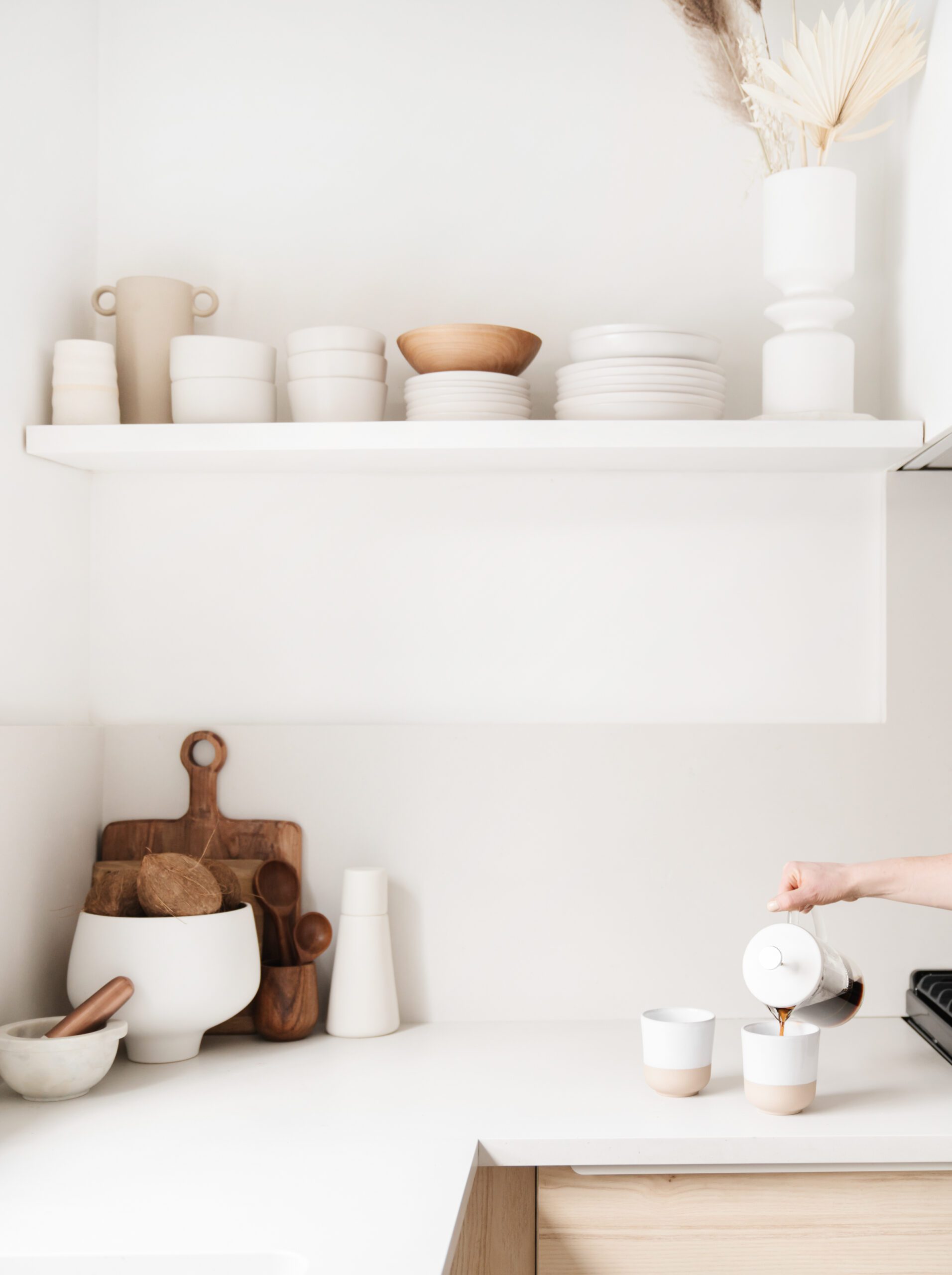 Minimalist kitchen with white walls, open shelf holding neutral dishes, and a person’s hand pouring coffee into cups on a clean counter with wooden cutting boards and kitchenware.