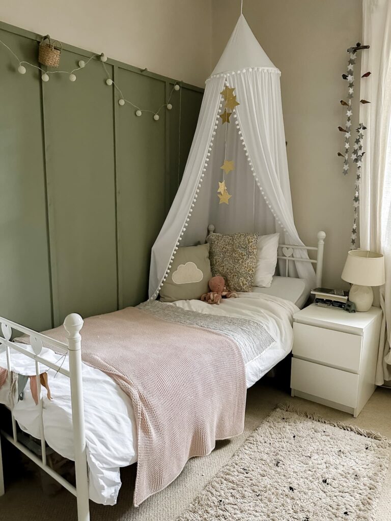 Cozy child’s bedroom with a white canopy and gold stars over a bed; pale green wall, plush toys, a pink blanket, bedside table, lamp, and string lights decorate the softly lit room.