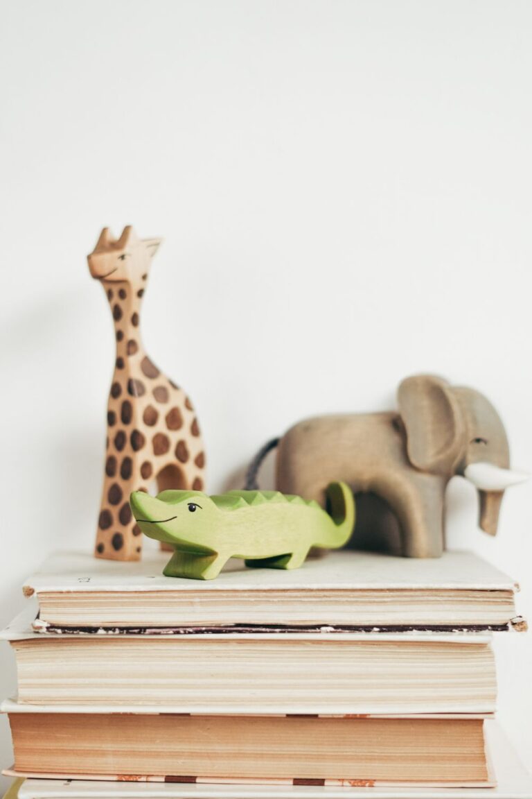 Three wooden animal toys—a giraffe, a green crocodile, and an elephant—are placed on top of a stack of books with a plain white background.