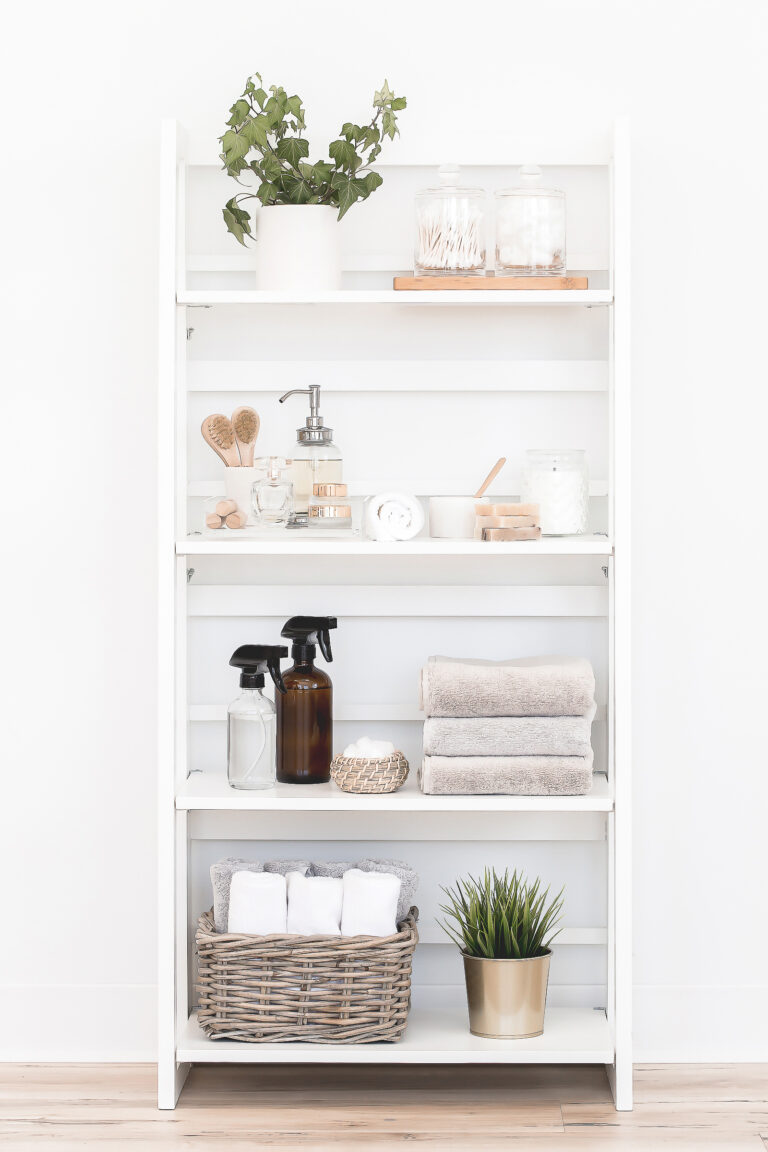White shelving unit with potted plant, glass jars, towels, bottles, basket with rolled towels, and decorative plant, arranged neatly against a white wall on a light wooden floor.