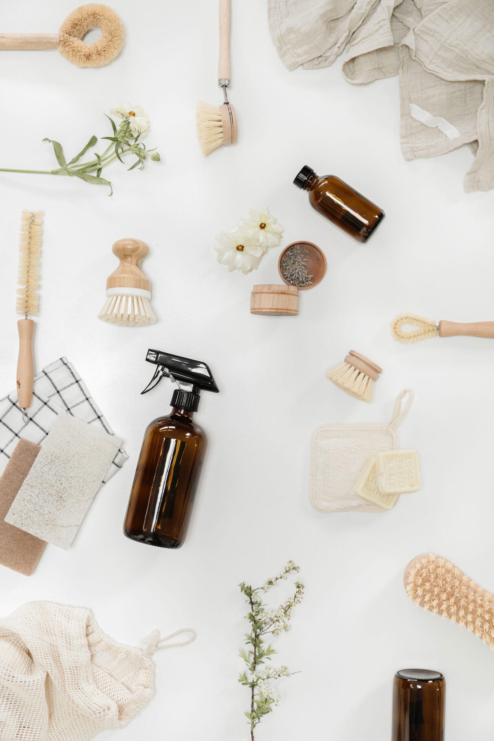 Brown glass bottles, wooden brushes, cloths, flowers, and eco-friendly cleaning items are arranged neatly on a white surface. The scene evokes a natural, sustainable cleaning theme.