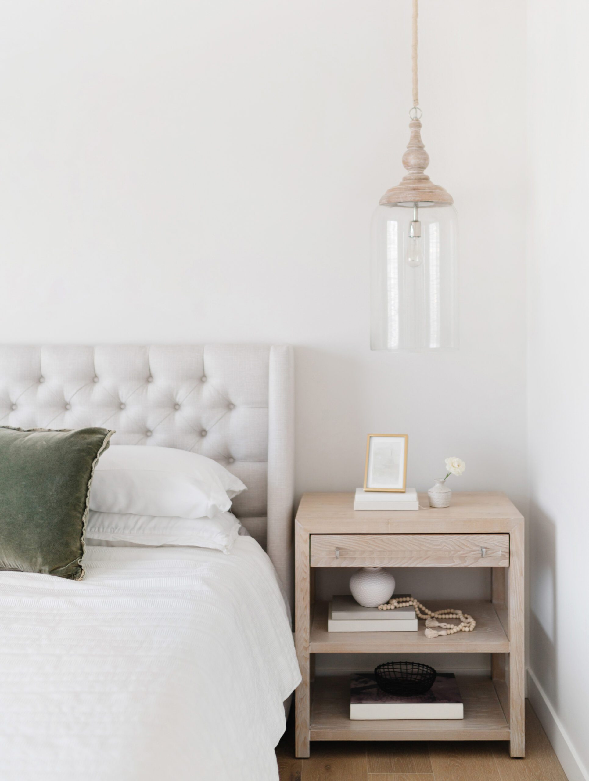 A minimalist bedroom with a tufted gray headboard, white bedding, a green pillow, a wooden nightstand with decor, and a glass pendant light hanging above. The decor is simple and modern.
