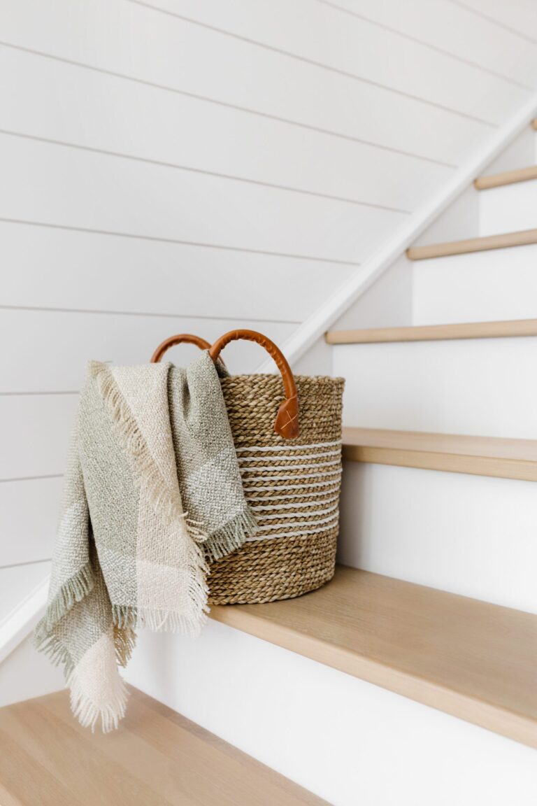 A woven basket with leather handles sits on wooden stairs, holding a fringed beige and white blanket. The background features white shiplap walls.
