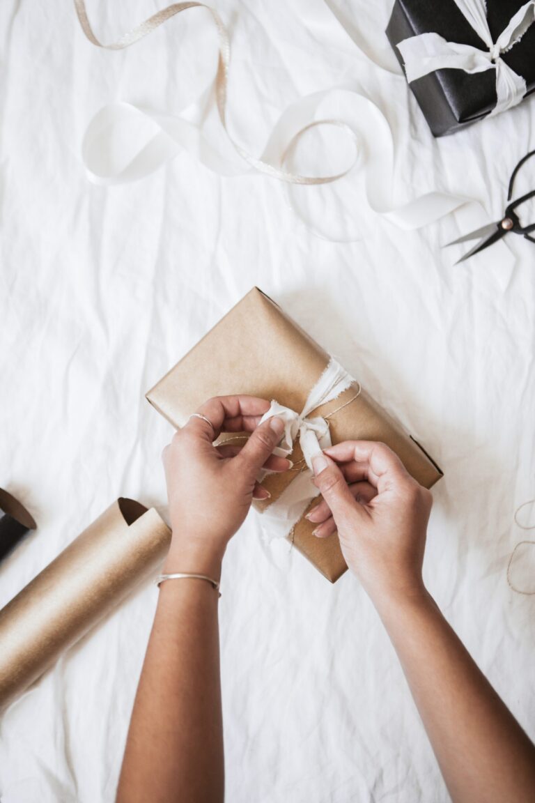 Hands tying a white ribbon around a brown gift box on a white surface, with wrapping paper, ribbon, and scissors nearby.