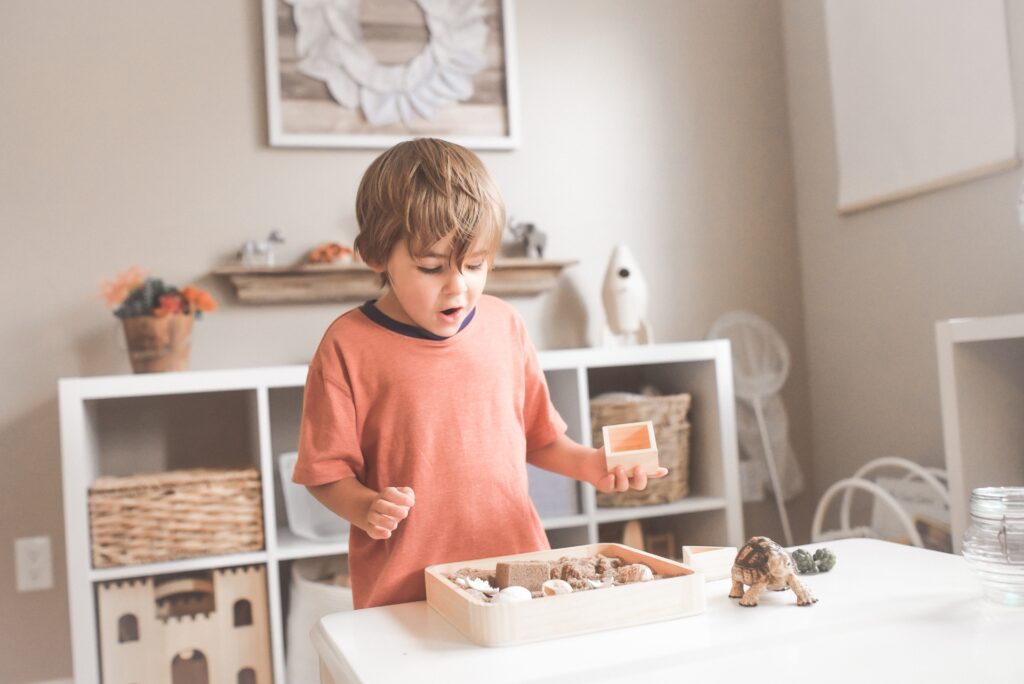 Young boy with brown hair wearing a peach shirt looks surprised while playing with a sensory box filled with sand and small toys in a light, tidy room.