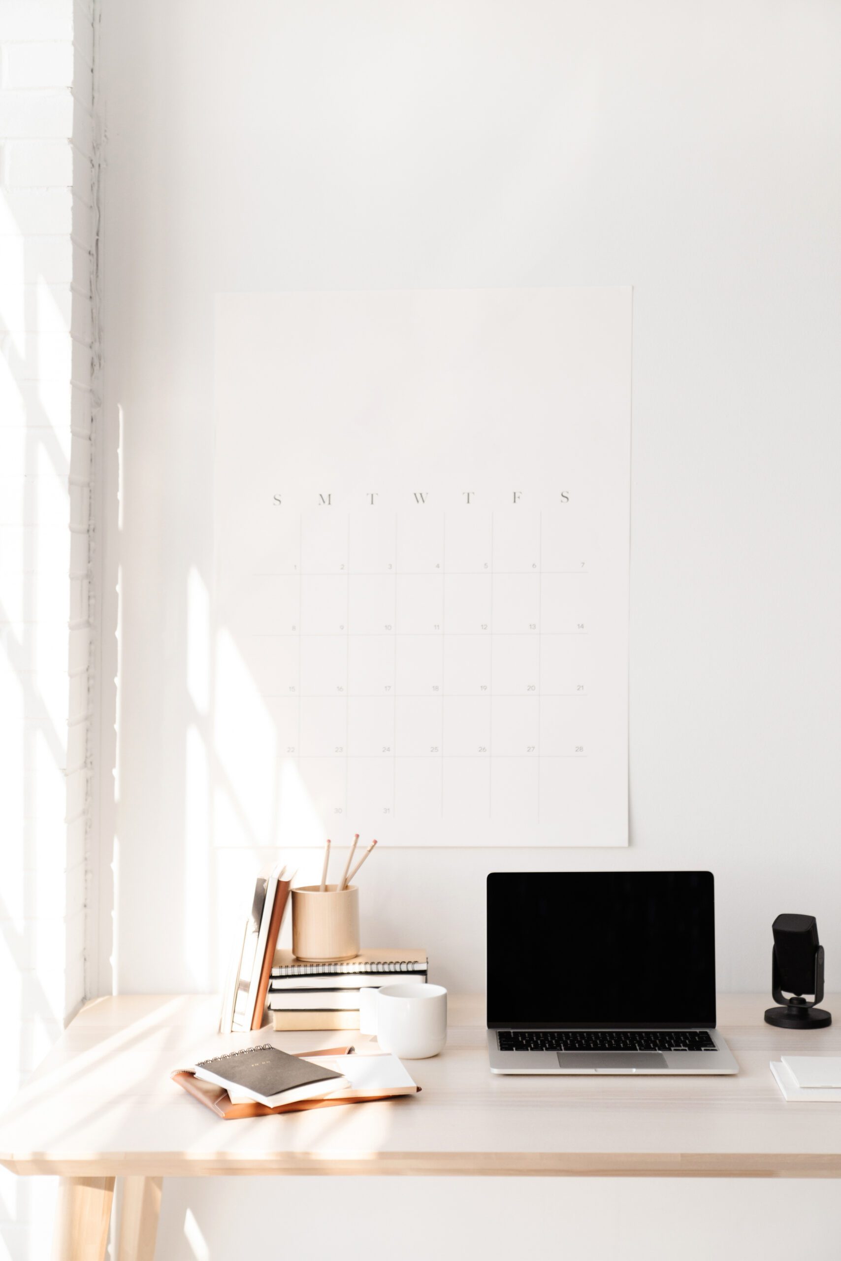 A minimalist desk with a laptop, notebooks, pencils, cup, and headphones sits by a sunlit wall with a large blank calendar hanging above.
