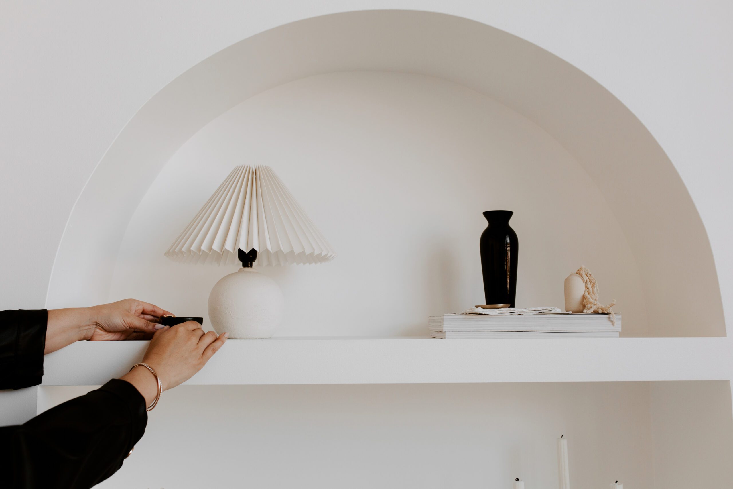 A person arranges a white shelf holding a pleated lamp, a black vase, a stack of books, and a seashell, set against a white arched wall.